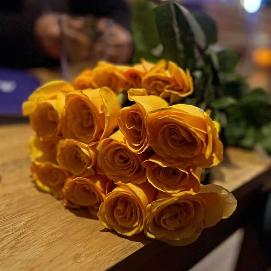Rosas amarillo dorado con reflejos naranja recostadas sobre una mesa de madera, con follaje verde detrás